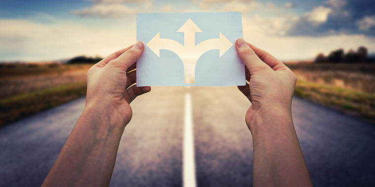 Zwei Hände halten eine Karte in der Hand, worauf drei Pfeile abgebildet sind. Ein Pfeil zeigt geradeaus, einer nach links und einer nach rechts. Im Hintergrund befindet sich eine Straße und ein sonniger Himmel