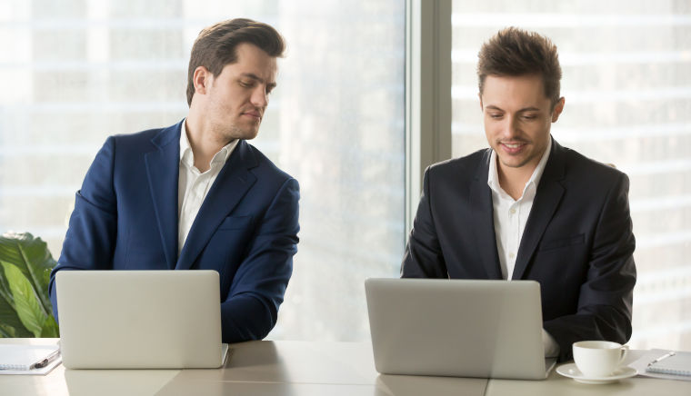 Zwei Geschäftsleute sitzen vor ihrem Laptop; einer schaut dem anderen über die Schulter, um Hilfe zu bekommen